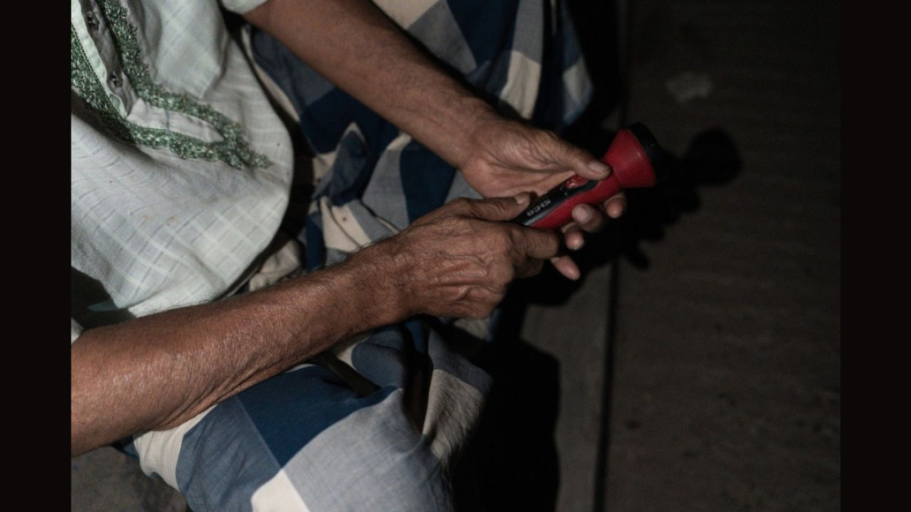 রাস্তার পাশে একা বসে হাতে লাল টর্চ ধরে বিদ্যুৎ ফেরার অপেক্ষা করছেন এক বৃদ্ধ। ছবি: সাদিক আল আশফাক