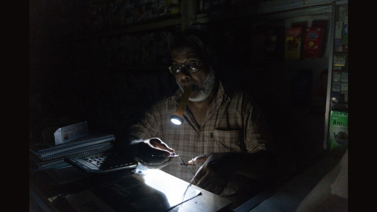 অন্ধকারের ভেতর ওষুধ কাটতে দাঁত দিয়ে টর্চলাইট চেপে ধরেছেন ফার্মেসির বিক্রেতা। ছবি: সাদিক আল আশফাক